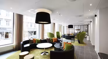 Modern lounge area with curved black sofas, green cushioned chairs, and orange pillows on green carpet. Large windows allow natural light to fill the room. Circular tables and overhead black lamps complete the cozy and stylish space.