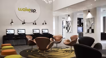 Modern hotel lobby with sleek chairs and round ottomans in earthy tones. Computer stations line the wall beneath a large wakeup aarhus logo. Stylish lighting and geometric mirrors enhance the contemporary design.