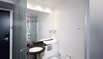 Modern bathroom with a large mirror above a dark countertop. It features a sink, toilet, and glass-enclosed shower. Towels are neatly folded near the sink, and a hairdryer is mounted on the wall. Ceiling lights illuminate the white walls.
