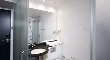 Modern bathroom with a large mirror above a dark countertop. It features a sink, toilet, and glass-enclosed shower. Towels are neatly folded near the sink, and a hairdryer is mounted on the wall. Ceiling lights illuminate the white walls.