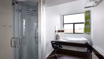 Compact hotel room featuring a glass-enclosed shower, a narrow bed by a window with green curtains, and a sleek black chair at a small desk. The room has white walls, wooden flooring, and a minimalist design.