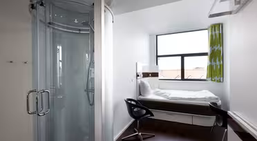 Compact hotel room featuring a glass-enclosed shower, a narrow bed by a window with green curtains, and a sleek black chair at a small desk. The room has white walls, wooden flooring, and a minimalist design.
