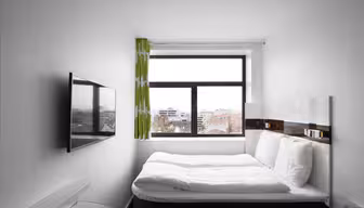 A compact, modern hotel room with a double bed, white bedding, and a wall-mounted TV. A large window with green curtains offers a cityscape view, featuring rooftops and buildings under a cloudy sky.