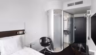 Minimalist hotel room with a white bed, small round glass table, and black chair. Features a modern, circular glass shower enclosure and a wall-mounted TV. The decor is sleek with black and white contrasts.
