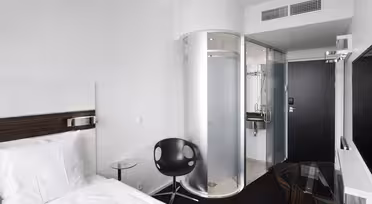 Minimalist hotel room with a white bed, small round glass table, and black chair. Features a modern, circular glass shower enclosure and a wall-mounted TV. The decor is sleek with black and white contrasts.