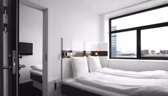 A minimalistic hotel room featuring a neatly made double bed with white bedding, a wall-mounted TV, large window with cityscape view, and modern furnishings. The room is bright and airy with a clean, contemporary design.