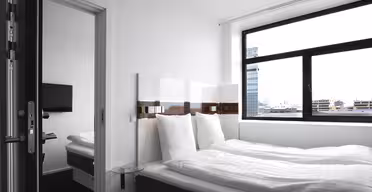 A minimalistic hotel room featuring a neatly made double bed with white bedding, a wall-mounted TV, large window with cityscape view, and modern furnishings. The room is bright and airy with a clean, contemporary design.