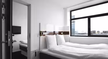 A minimalistic hotel room featuring a neatly made double bed with white bedding, a wall-mounted TV, large window with cityscape view, and modern furnishings. The room is bright and airy with a clean, contemporary design.