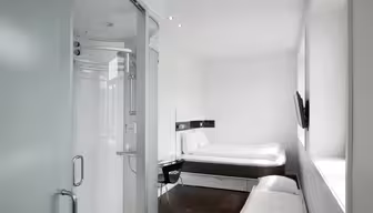 A modern, small hotel room with a minimalist design. It features a double bed, mounted TV, a chair by a desk, and a partially visible glass-encased shower. The room has white walls and dark wood flooring, creating a clean, bright atmosphere.