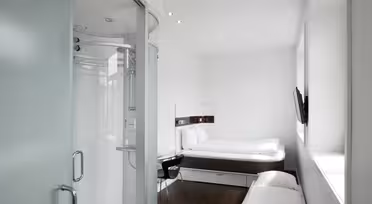 A modern, small hotel room with a minimalist design. It features a double bed, mounted TV, a chair by a desk, and a partially visible glass-encased shower. The room has white walls and dark wood flooring, creating a clean, bright atmosphere.