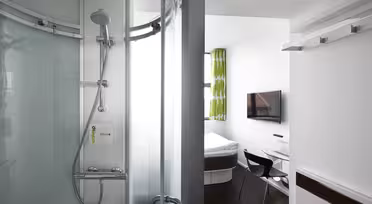 A compact, modern hotel room featuring a corner shower with frosted glass doors and a wall-mounted showerhead. The room includes a small bed, desk, chair, flat-screen TV, and a window with green patterned curtains.