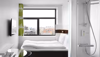 A compact hotel room features a neatly made bed, a wall-mounted TV, a glass-top table, and a shower with clear glass doors. A large window with green curtains offers a view of city rooftops under a cloudy sky.
