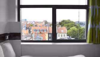 View from a hotel room window showing a cityscape with a mix of red-roofed buildings and greenery. Partial view of a bed with white sheets in the foreground. Overcast sky visible.