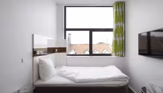 A small, modern bedroom with a neatly made bed, white bedding, and a large window with green and white curtains. Theres a wall-mounted TV on the right and a glass side table beside the bed. Roofs of neighboring buildings are visible through the window.