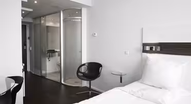 Modern hotel room with a single bed, white bedding, and a dark wood floor. The room features a sleek black chair, a round glass side table, and a glass-enclosed bathroom with a toilet and shower. The walls are white, enhancing the minimalist style.