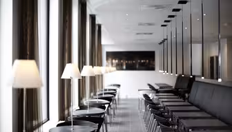 A modern cafe interior with a row of black tables and chairs alongside a long window. Each table has a small white lamp. The room features a sleek design with high ceilings and mirrored walls.