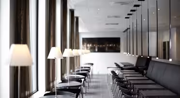 A modern cafe interior with a row of black tables and chairs alongside a long window. Each table has a small white lamp. The room features a sleek design with high ceilings and mirrored walls.