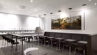A modern cafe interior with a row of small tables and chairs along a wall. Barstools line a high counter on the left. The walls are white with minimal decor, featuring a large photo of a garden scene. Ceiling lights provide illumination.