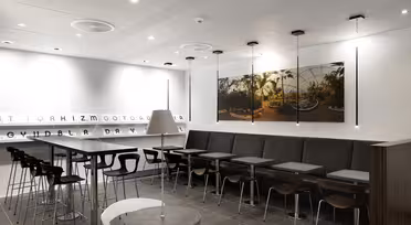 A modern cafe interior with a row of small tables and chairs along a wall. Barstools line a high counter on the left. The walls are white with minimal decor, featuring a large photo of a garden scene. Ceiling lights provide illumination.