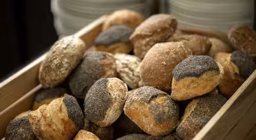 A wooden crate filled with various freshly baked bread rolls, some topped with poppy seeds. Stacks of plates are visible in the blurred background.