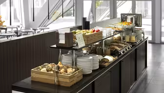 A modern buffet setup featuring a variety of breakfast items on a long counter. Includes baskets of bread rolls, plates, fresh fruits, and a toaster. Wooden accents and large windows visible in the background.
