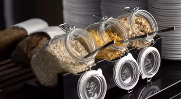 Three glass jars are tilted forward on a black rack, containing oats, cornflakes, and granola. Each jar has a spoon resting inside. Stacked white plates and a loaf of bread are visible in the background.