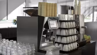 A modern coffee machine with a digital interface is surrounded by stacked white cups and rows of plastic glasses. Nearby are paper cups and a display of assorted tea bags in a well-lit café setting.