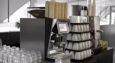A modern coffee machine with a digital interface is surrounded by stacked white cups and rows of plastic glasses. Nearby are paper cups and a display of assorted tea bags in a well-lit café setting.