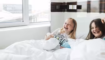 Two young girls laughing and lying in bed near a large window with a city view. Both are covered with white blankets. The girl on the left wears a graphic t-shirt, and the scene is filled with natural light, creating a cheerful atmosphere.