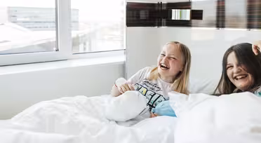 Two young girls laughing and lying in bed near a large window with a city view. Both are covered with white blankets. The girl on the left wears a graphic t-shirt, and the scene is filled with natural light, creating a cheerful atmosphere.