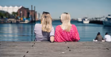 Two people with long hair sit on a wooden dock facing a large body of water. One wears a purple top and the other a pink top. In the background, there are buildings, boats, and a clear blue sky.