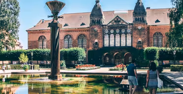 A couple walks towards a historic brick building with large arched windows and ornate details. In the foreground, a fountain with a tall, vertical sculpture is surrounded by greenery and colorful flowers.