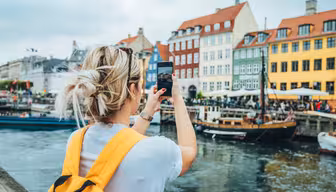 A person with blond hair and a yellow backpack is taking a photo of colorful buildings along a canal. Boats are docked in the water, and people are visible on the waterfront. The sky is overcast.