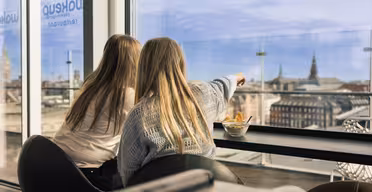 Two people with long hair sit indoors by a window, overlooking a cityscape. One points outside, while the other watches. A bowl of snacks is on the table in front of them. The scene conveys a relaxed, observational moment.