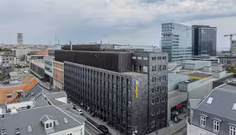 Aerial view of an urban landscape featuring a modern, dark-colored hotel labeled Wakeup in a busy city area. Surrounding buildings vary in style, with a mix of older and contemporary architecture. The sky is overcast, adding a gray tone to the scene.