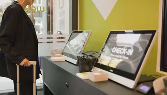A person stands at a check-in counter, holding a suitcase handle. Two computer screens display CHECK-IN in a bright, modern setting. The walls are green, and office supplies are neatly arranged on the counter.