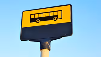 A bus stop sign with a yellow background and a black silhouette of a bus. Its mounted on a post, with a blue sky in the background.