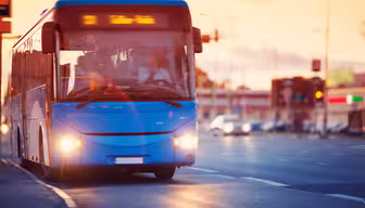 A blue city bus drives along a road at sunset, its headlights on and destination displayed on the front. The sky is orange, and buildings with blurred lights are in the background, with the road partly illuminated by streetlights.