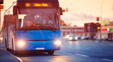 A blue city bus drives along a road at sunset, its headlights on and destination displayed on the front. The sky is orange, and buildings with blurred lights are in the background, with the road partly illuminated by streetlights.