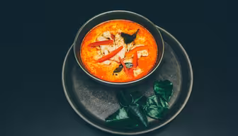 A bowl of red curry with tofu, garnished with red chili slices and herbs, is placed on a dark plate. Two sprigs of green leaves are beside the bowl. The dish is on a dark surface.