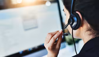 Woman wearing a headset sits at a computer, focusing on the screen. She holds the microphone near her mouth, suggesting she is engaged in a call or providing customer support. The screen displays blurred text.