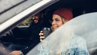A woman wearing a red beanie and plaid shirt smiles while holding a coffee cup inside a car. A man wearing a dark jacket sits beside her in the drivers seat, both appear cheerful as they look out the window.