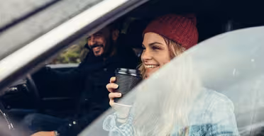 A woman wearing a red beanie and plaid shirt smiles while holding a coffee cup inside a car. A man wearing a dark jacket sits beside her in the drivers seat, both appear cheerful as they look out the window.