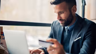 A man sits at a table by the window, concentrating on his laptop. Hes wearing a gray suit and holding a credit card, suggesting online shopping or a financial transaction. The background is slightly blurred, emphasizing a bright, natural atmosphere.