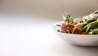 A white ceramic bowl filled with a fresh salad. The salad includes greens, sliced red onions, lentils, and croutons, all against a softly blurred light background.