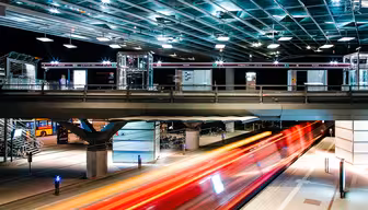 A brightly lit, modern train station at night with a sleek, glass-covered platform. A train rushes by, creating streaks of red and orange lights. The station features metal structures and a pedestrian walkway above.