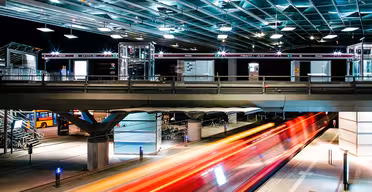 A brightly lit, modern train station at night with a sleek, glass-covered platform. A train rushes by, creating streaks of red and orange lights. The station features metal structures and a pedestrian walkway above.