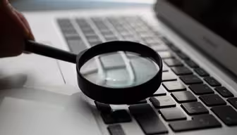 A magnifying glass hovers over the keyboard of an open laptop, highlighting the keys in focus. Sunlight creates a reflection on the laptop surface.