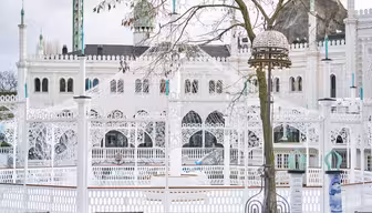 A winter scene at Tivoli Gardens in Copenhagen, featuring the white Moorish-style Nimb Hotel. Intricate arches and domes are adorned with snow. Bare tree branches partially obscure the view. A decorative lamp stands in the foreground.