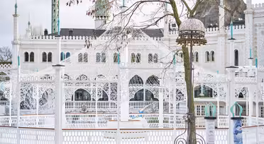 A winter scene at Tivoli Gardens in Copenhagen, featuring the white Moorish-style Nimb Hotel. Intricate arches and domes are adorned with snow. Bare tree branches partially obscure the view. A decorative lamp stands in the foreground.